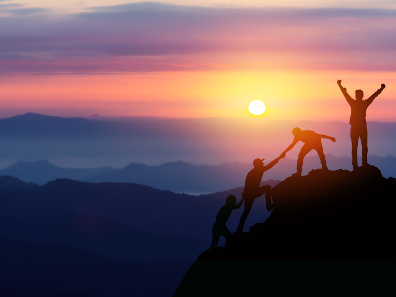 Group of friends climb mountain at sunrise while one friend celebrates at the top.