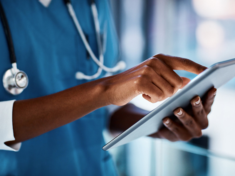 Lower half of African-American man wearing blue scrubs and a stethoscope scrolling through a document on a tablet.