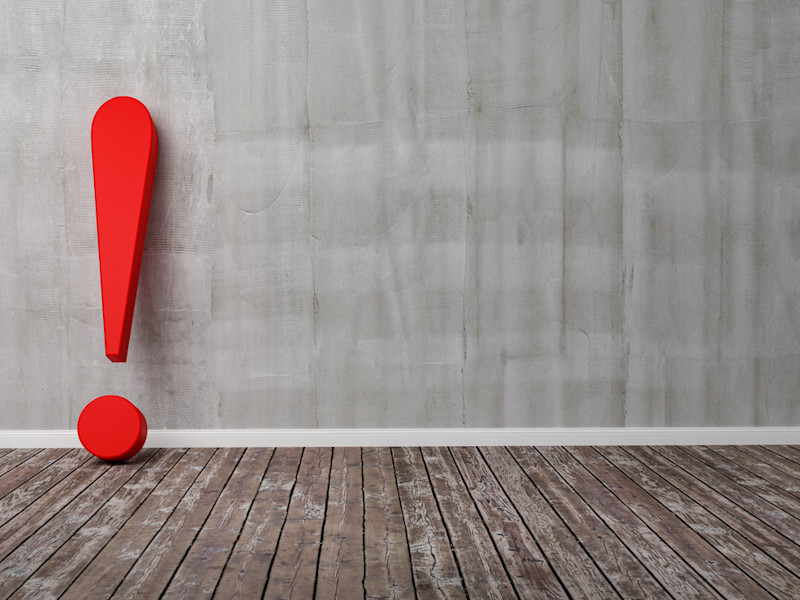 Red exclamation point standing on a wooden floor and resting against a gray wall.