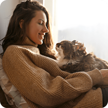 A smiling woman who is looking so relieved that she acquired pet insurance for her beautiful long-haired cat sitting on her lap and lovingly gazing at her.