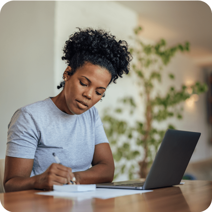 A focused student sits at a desk with a laptop, thoughtfully writing notes while working on their scholarship application in a calm, plant-filled study space