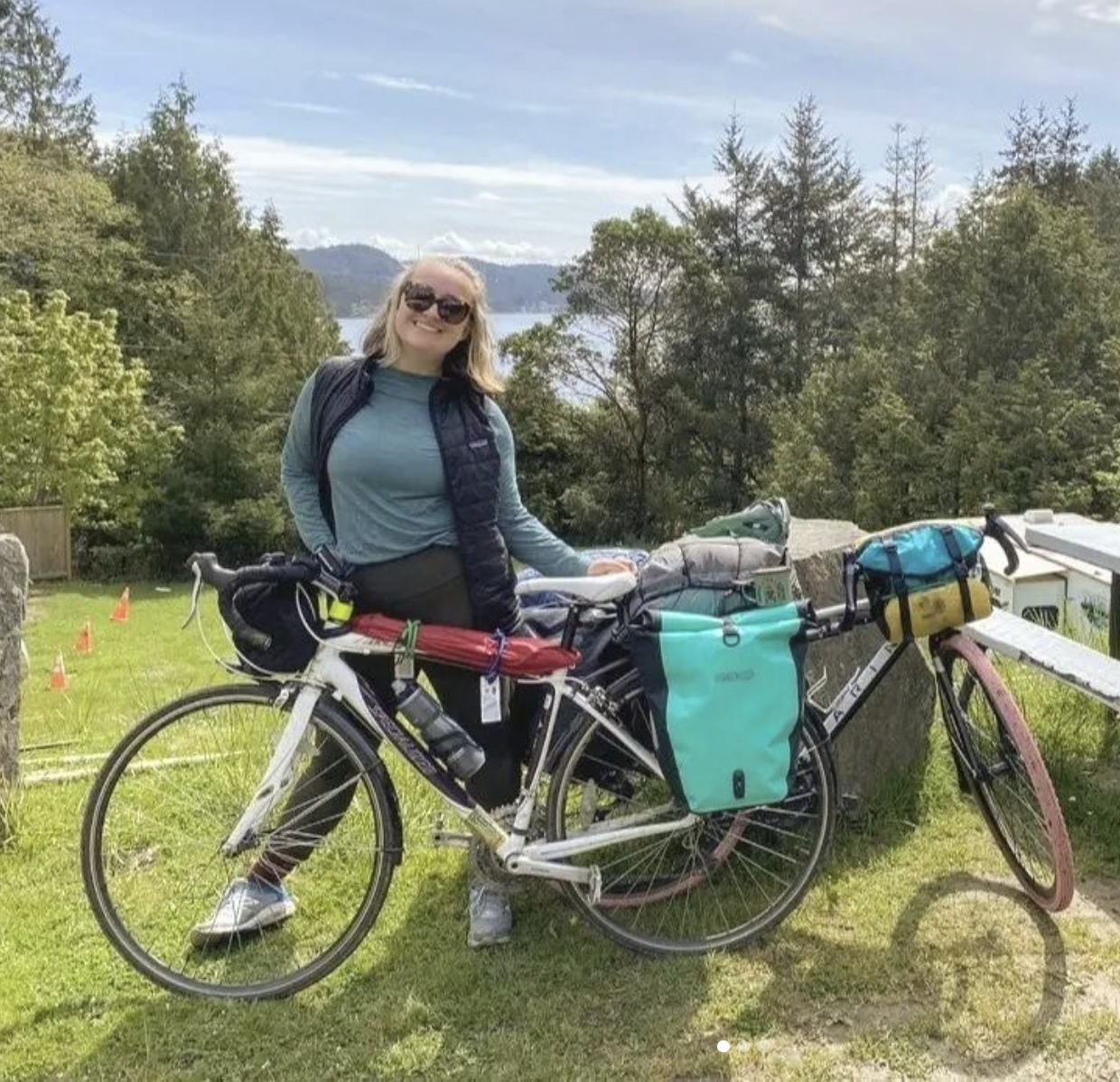 An image of Wren smiling and standing behind their loaded touring bike, against a background of trees, the ocean, and Vancouver island.
