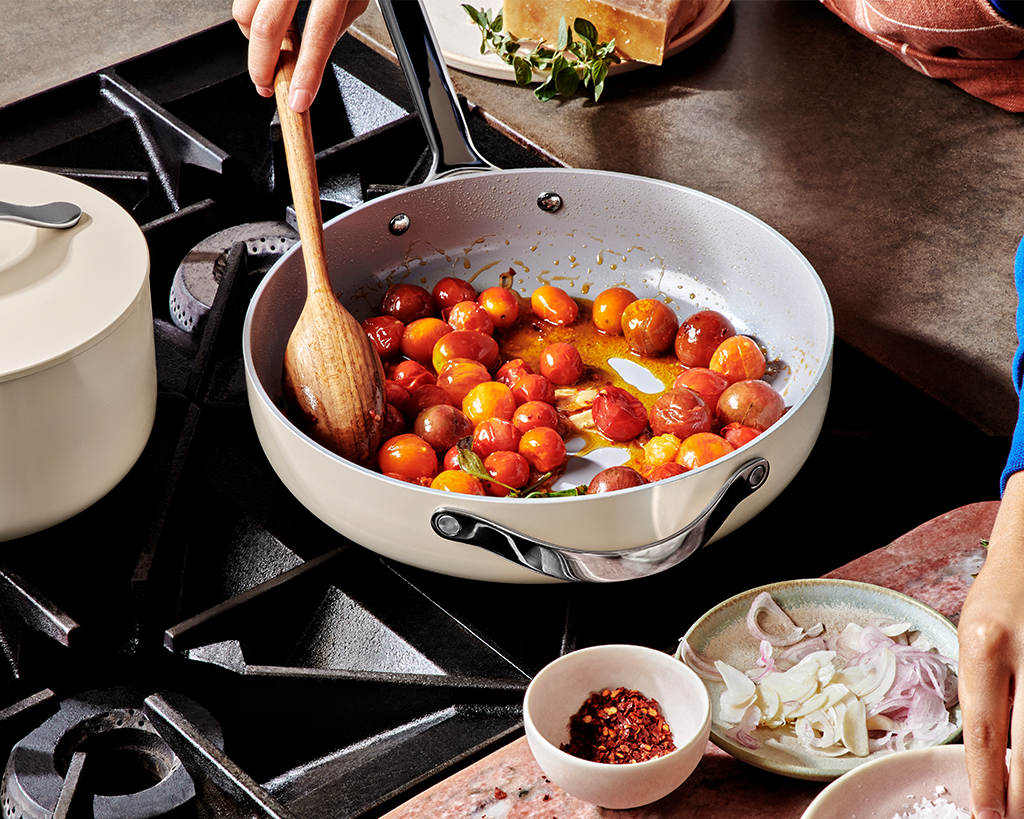 A white ceramic non-stick sauté pan and a wooden ladle being used to cook tomatoes on a stove top