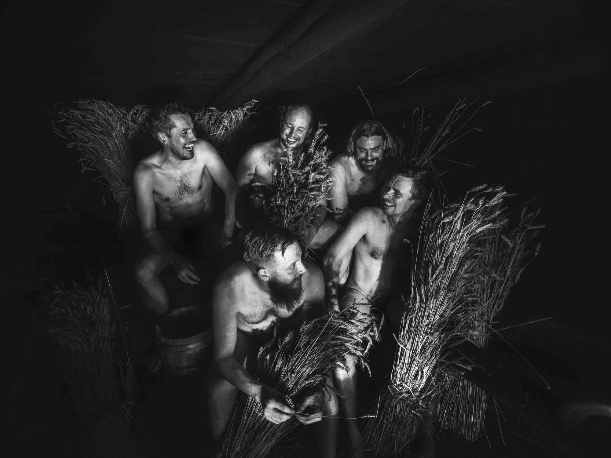 A black and white image of the five Kyrö founders sitting in a midsummer sauna surrounded by bushels of rye. 