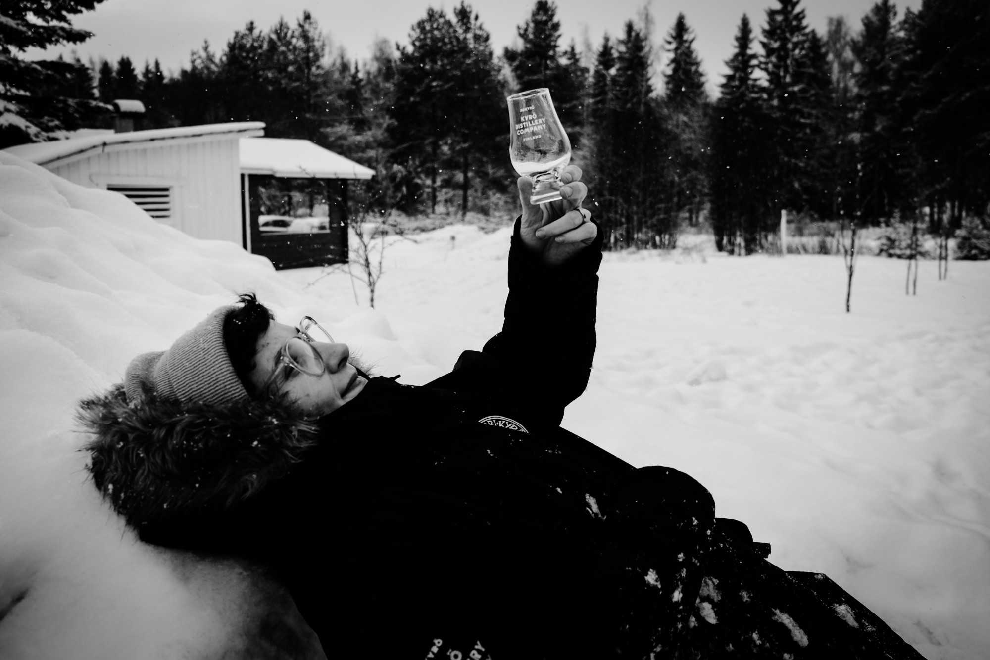 Woman laying in snow and staring at an empty Glencairn whisky tasting glass.