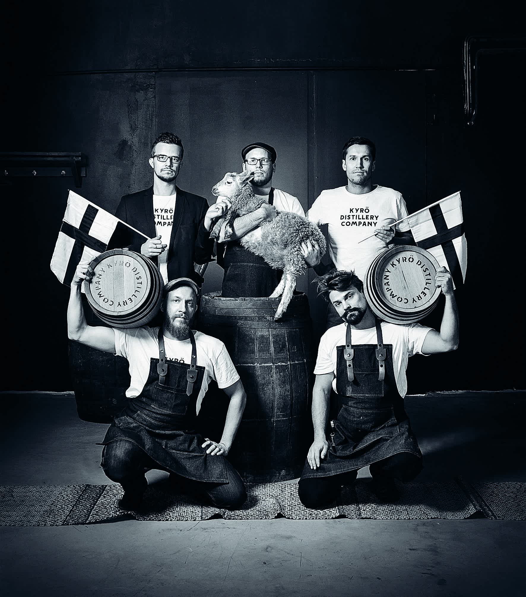 Black and white image: the five founders of the Kyrö Distillery Company in a group picture, two holding barrels, two holding Finnish flags, one holding a lamb. 