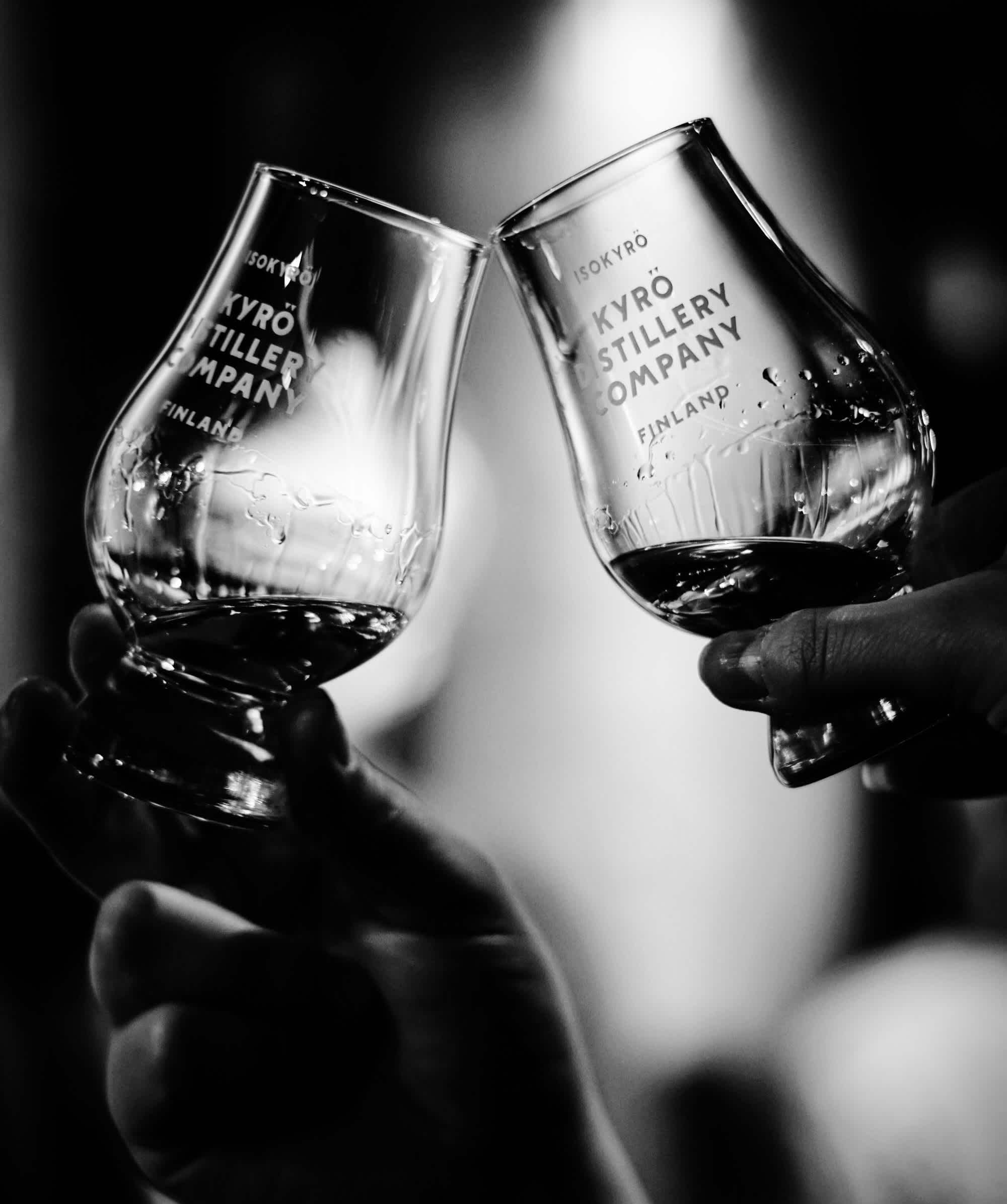 Black and white image of two Kyrö-branded Glencairn tasting glasses filled with whisky cheersing each other. 