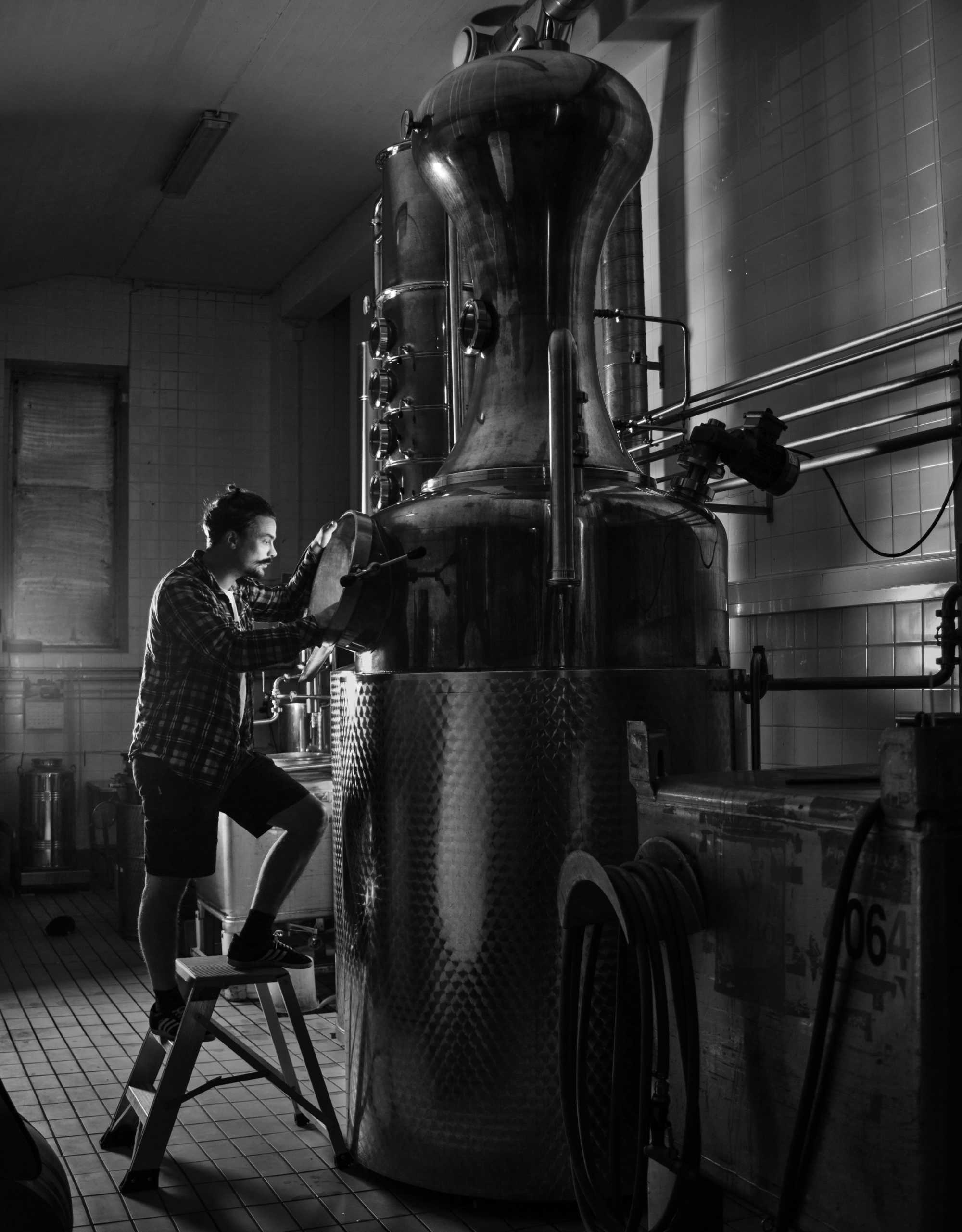 Black and white image of head distiller Kalle Vallkonnen standing on a ladder, peering into a distilling pot. 