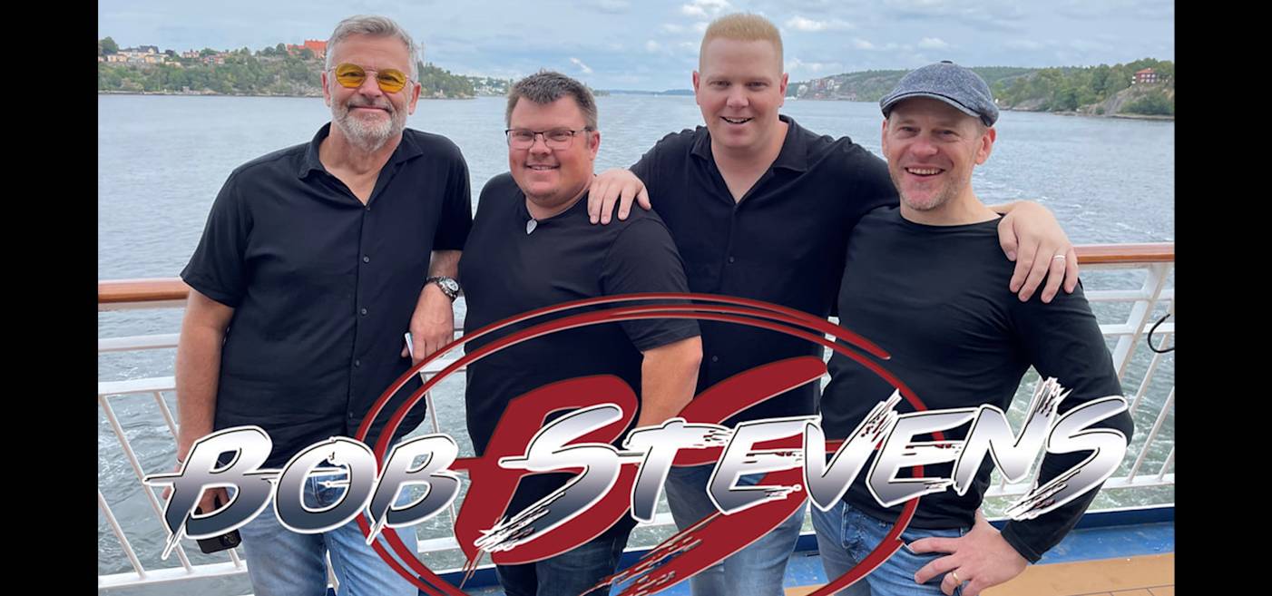 Fyra män i fritidskläder står tillsammans på ett båtdäck med vatten och hus i bakgrunden. Texten över bilden lyder "Bob Stevens.