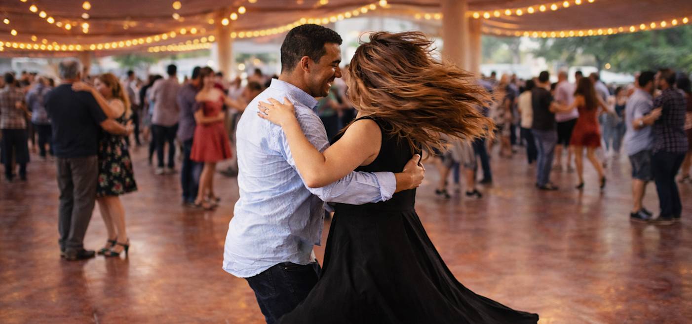 En man och en kvinna dansar tillsammans i en väl upplyst utomhuslokal med mycket folk, ljusslingor i taket och andra dansande människor i bakgrunden.