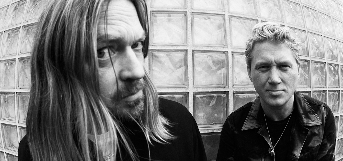 Two men are posed in front of a glass block wall. One has long hair and a beard; the other has short hair. Both wear casual jackets and look directly at the camera. Image is in black and white.