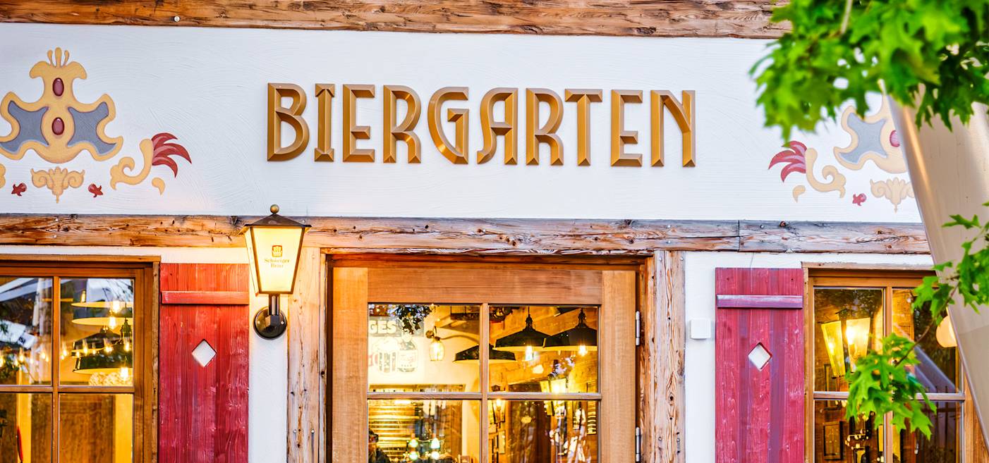 Exterior of a building with a sign reading "Biergarten" above wooden doors and decorative trim, illuminated by a wall-mounted lamp.