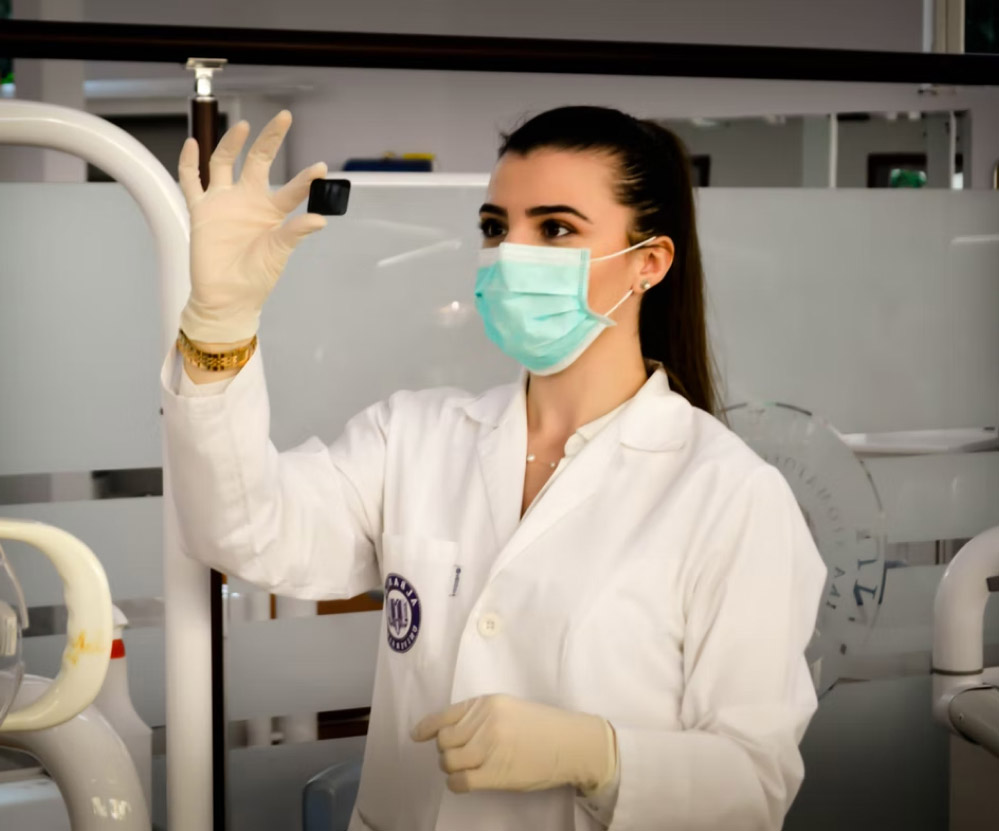 Femme qui porte une blouse de laboratoire, un masque et des gants, tenant un petit objet