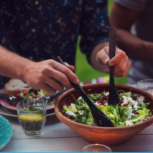 A person tossing a salad with serving utensils, a glass of water and food in background.
