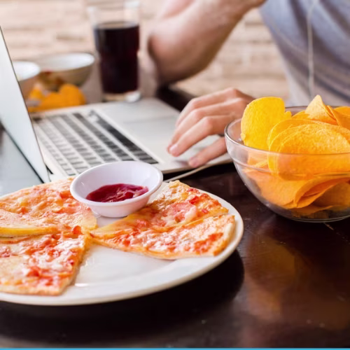 A person working on a laptop while eating pizza, chips and beverage.