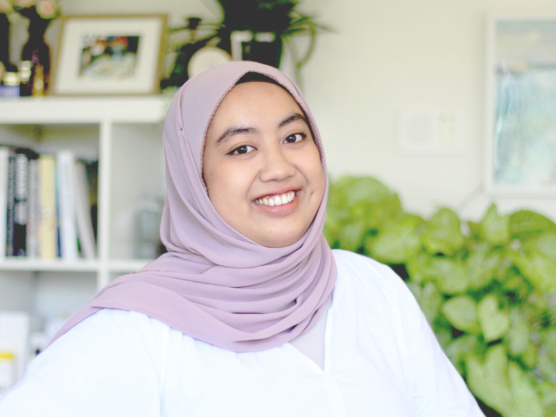 A photo of Farhana working from home. There's a book shelf and plants in the background.