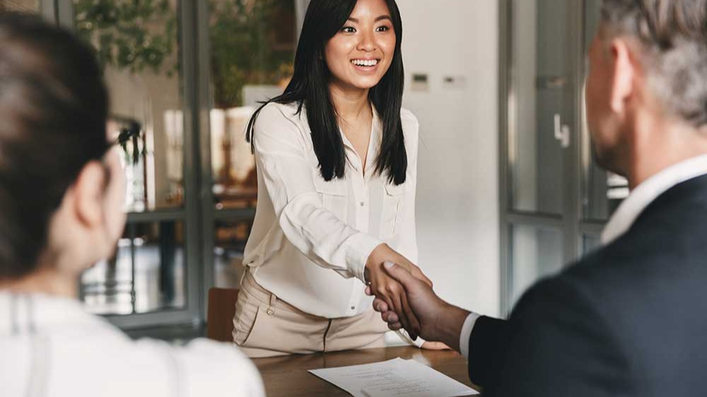 image from back of two employers sitting in office and shaking hand of young woman