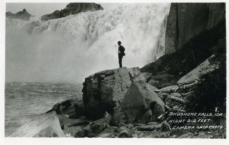 A man stands on a large rock near a waterfall. 