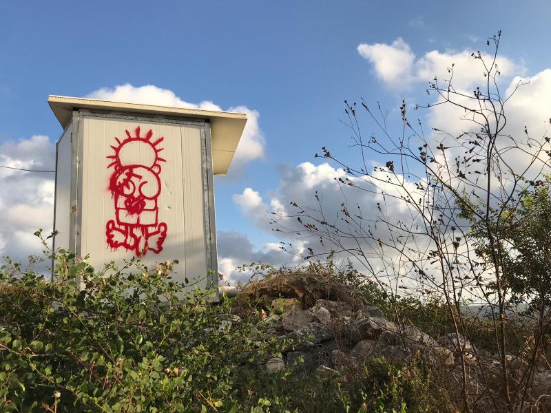 Red graffiti of handala, a Palestinian symbol of resistance created by Naji el-Ali, of a 10-year-old boy with his back turned on the viewer and clasped hands behind his back. There is a blue sky and greenery surrounding the graffiti'd wall.