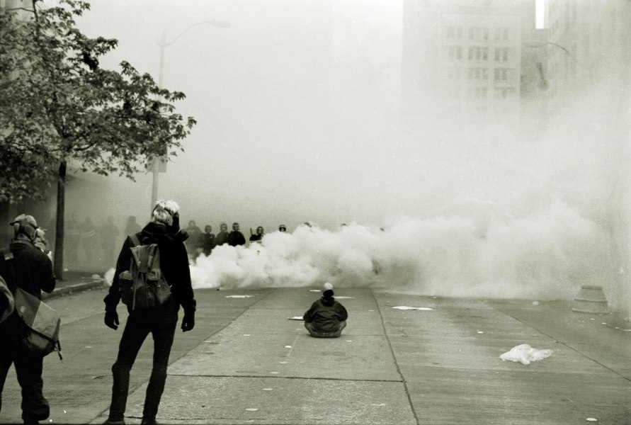 A photo of a few protesters facing a line of riot police and a cloud of tear gas. 
