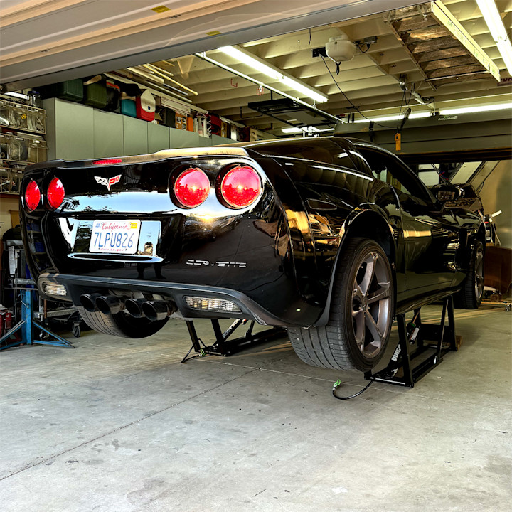 Black Chevrolet Corvette with its hood open, raised on a QuickJack portable car lift in a home garage.