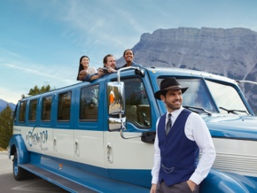 Three tourists poke their heads out of the open roof of the touring vehicle to admire the mountain views. The driver-guide, wearing vintage clothes, stands beside the vehicle.