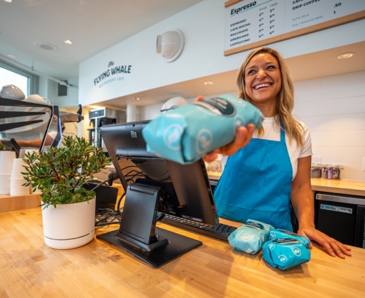 A barista at the Flying Whale Cafe smiles as she sells a customer a fresh sandwich wrapped in paper.