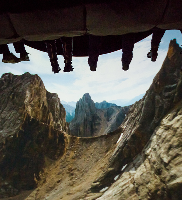 People enjoying Flyover Canada appear to soar through a mountain canyon. Their dangling feet are seen from behind as the people sit in the flying ride. 