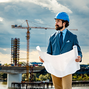 A construction professional wearing a blue hard hat holds architectural plans while inspecting a building site with cranes in the background.