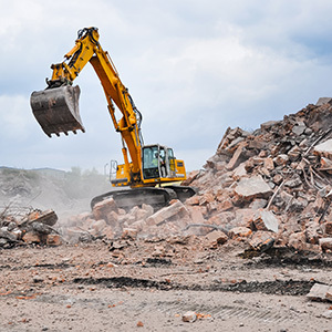 A large yellow excavator uses its bucket to break and move piles of concrete and debris at an active demolition site.