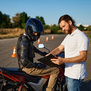 A motorcyclist wearing protective gear sits on a motorcycle while an instructor reviews information on a clipboard during a riding lesson.