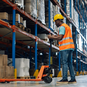 A warehouse worker wearing a high-visibility safety vest and hard hat uses a manual pallet jack to move materials through a warehouse aisle lined with tall storage racks and stacked boxes.