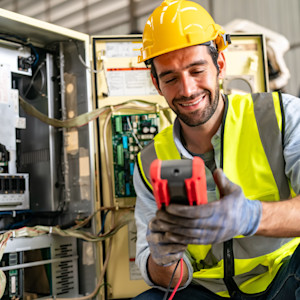A professional electrician wearing a yellow hard hat, high-visibility safety vest and work gloves uses a digital multimeter to test wiring inside an open electrical control panel.
