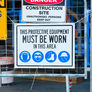 A safety sign mounted on a construction site fence states that protective equipment must be worn in the area, with blue icons showing required hearing protection, safety glasses, work boots and hard hats.