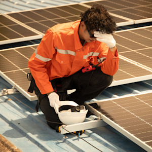 An overheated construction worker crouches on a metal roof with solar panels.