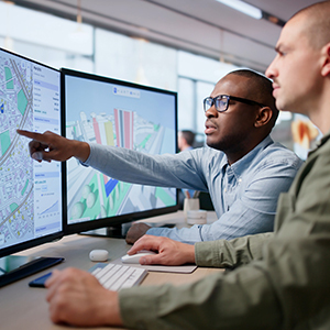 Two professionals analyze detailed city maps and planning data displayed across multiple computer screens.