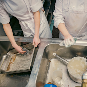 Two employees scrub cookware in a stainless steel multi-compartment sink in a commercial kitchen.