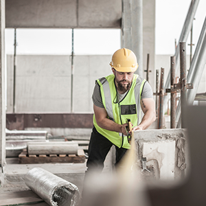 A construction worker wearing a hard hat and high-visibility vest uses a power tool to cut or break concrete inside a building under construction.
