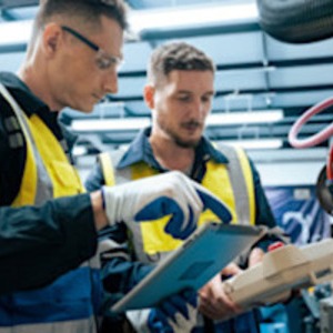 Two maintenance technicians troubleshoot equipment at an industrial control panel, one demonstrating the controls to the other on a factory floor.