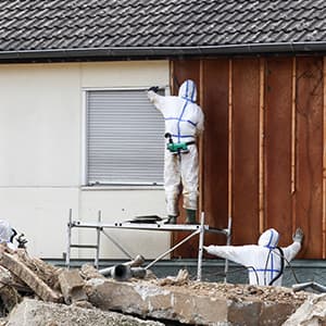 Three workers wearing full protective suits and respirators inspect the exterior of a building during renovation or hazardous material removal.