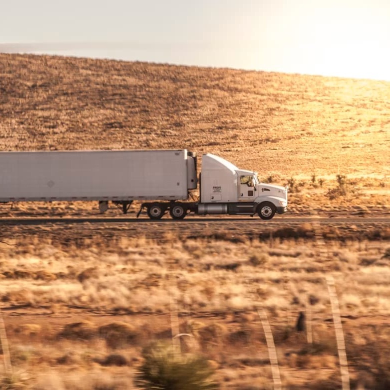 A white commercial semi-truck transports a long enclosed trailer along a highway surrounded by dry, sunlit hills.