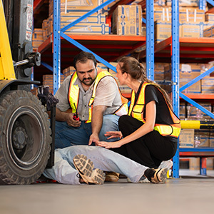 Two warehouse employees wearing high-visibility safety vests kneel beside an injured coworker lying on the floor near a forklift, appearing to assess the situation and provide help.