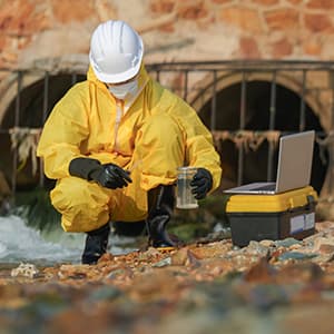 A worker wearing a yellow protective suit, helmet, mask, and gloves collects a water sample near a drainage area while using a laptop and sampling equipment.