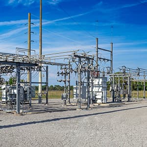 An outdoor electrical substation contains transformers, power lines, metal structures, and other high-voltage equipment under a clear blue sky.