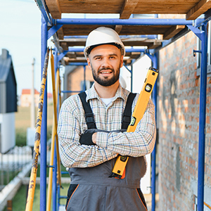 A construction worker wearing a hard hat and work overalls stands near scaffolding while holding a level.