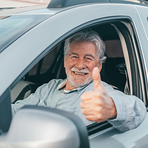 An older man with gray hair and a beard smiles warmly while sitting in the driver’s seat of a car, giving a thumbs-up gesture out the window to show confidence and positivity.