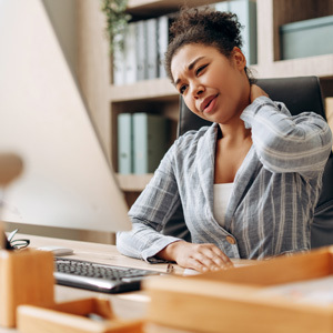 A woman sitting at her office desk in front of a computer screen holds her neck with a pained expression.