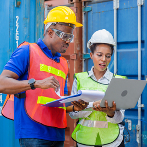 Two construction workers wearing hard hats, safety glasses and high-visibility vests review paperwork and a laptop near shipping containers at a worksite.