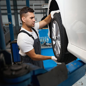 A mechanic expects a wheel on a white car on a lift in a shop.