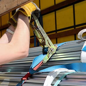 A worker wearing protective gloves uses a ratchet tensioner to secure metal pipes with heavy-duty straps at a construction site.